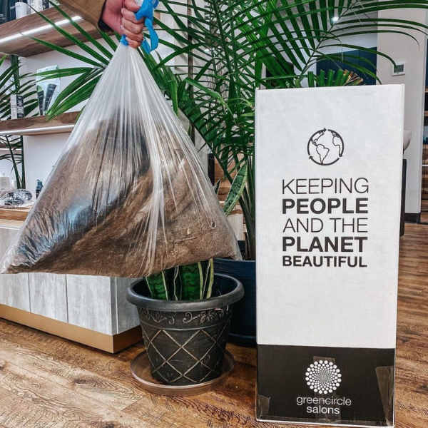 A hand holding a trash bag full of hair in salon with plants in the background, next to a GreenCircle salon recycle box.