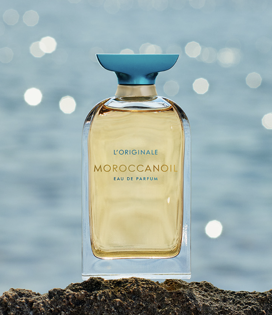 Moroccanoil's fine fragrance perfume bottle has a golden liquid and teal cap up against a beachy background