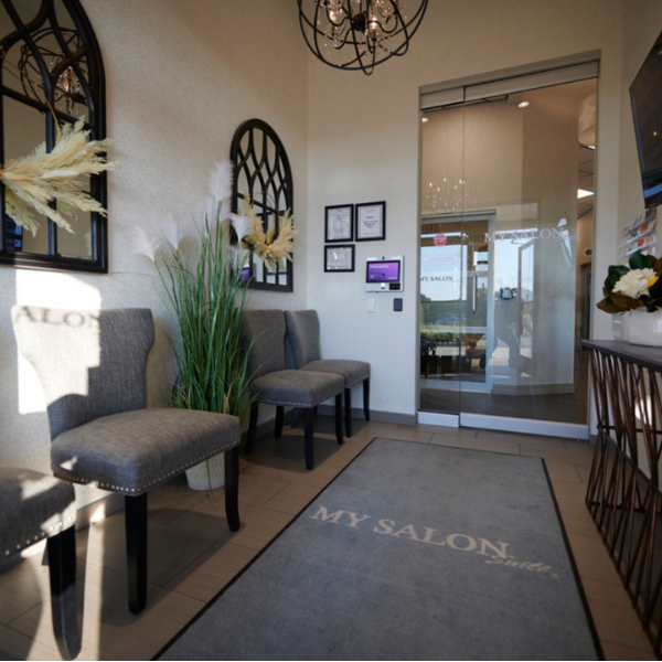 Elegant MY SALON Suite® lobby with gray chairs, decorative mirrors, and a welcoming entrance featuring branded décor and soft lighting.