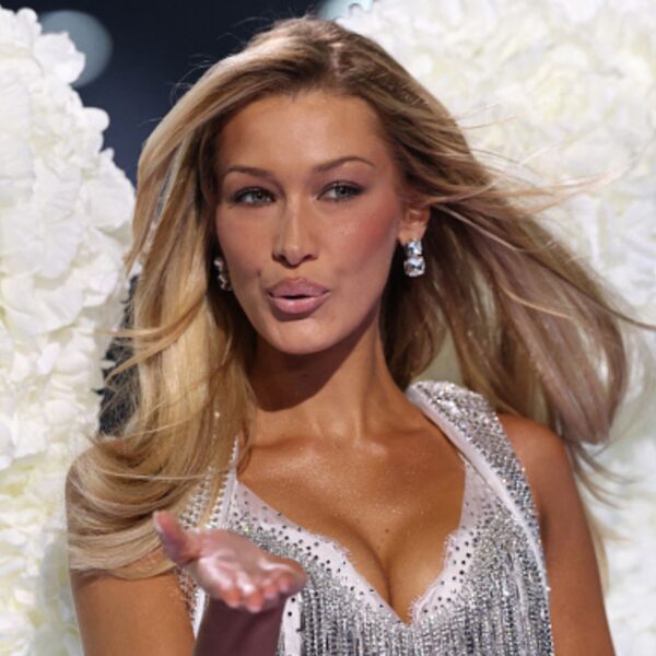 Bella Hadid with long, voluminous blonde hair styled in soft waves is seen on the runway, blowing a kiss while wearing a sparkly silver outfit with feathered white wings in the background.