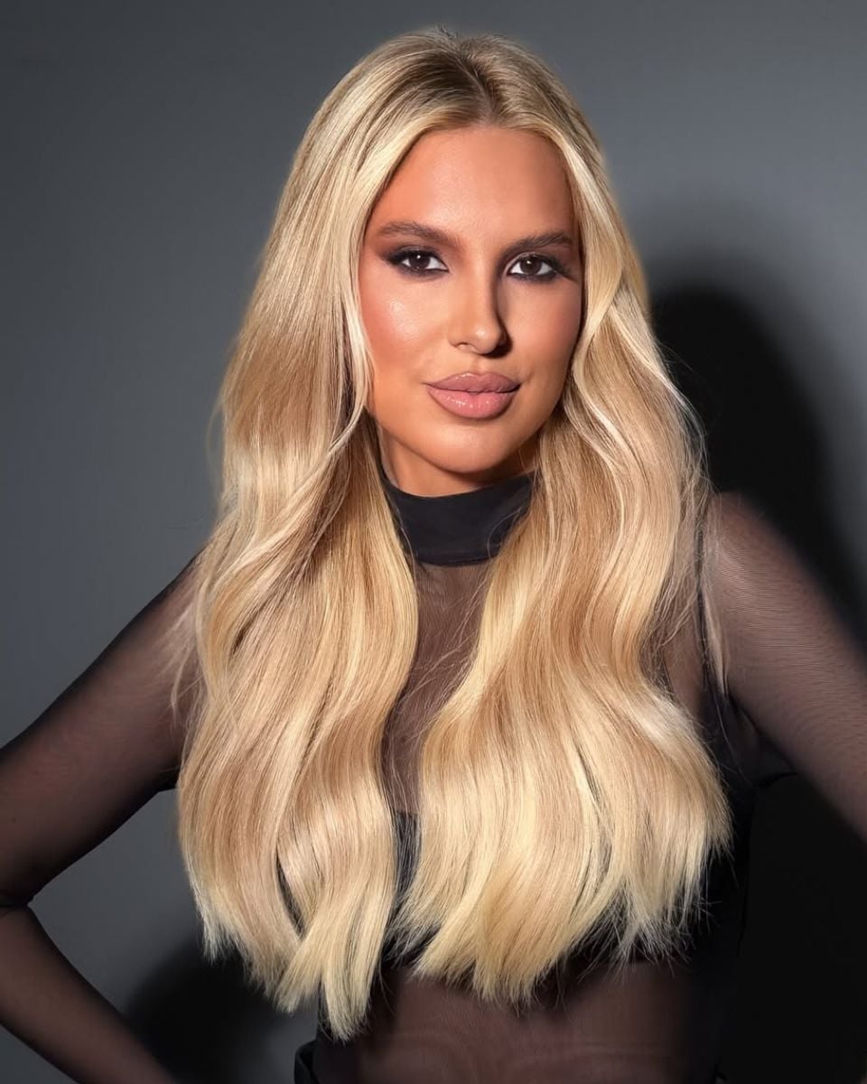 Portrait of long champagne blonde hair with soft beige dimension, a seamless root blend and polished waves styled over a sheer black top.